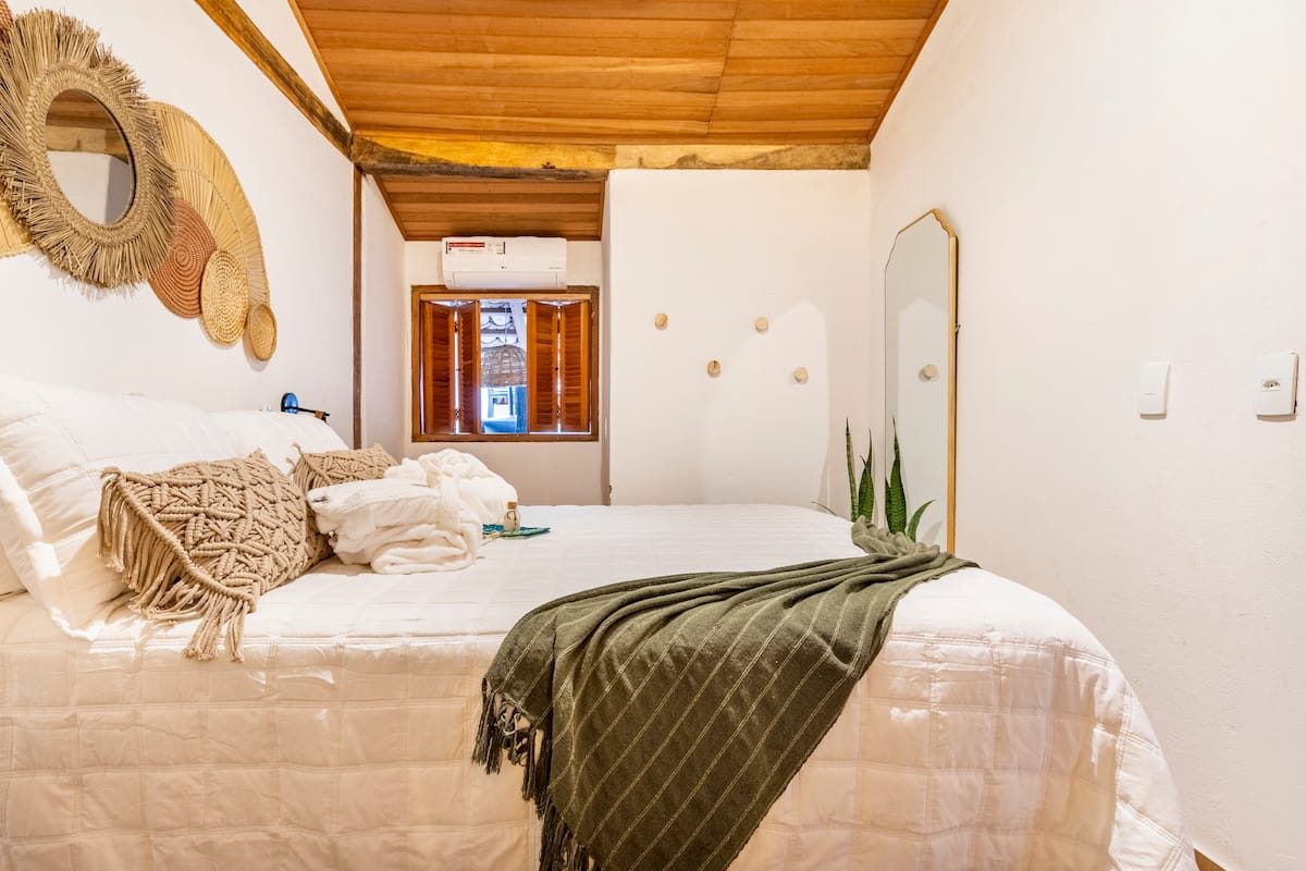 Quarto claro da Casa Boutique com cama branca, manta verde e espelho redondo de palha