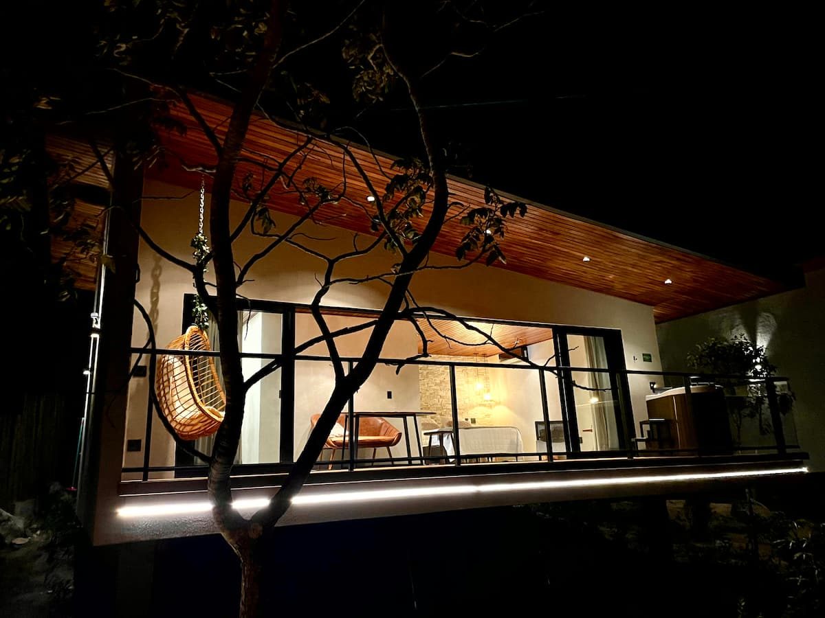 Casa Jade iluminada à noite, com cadeira suspensa na varanda e o interior visto pelo vidro