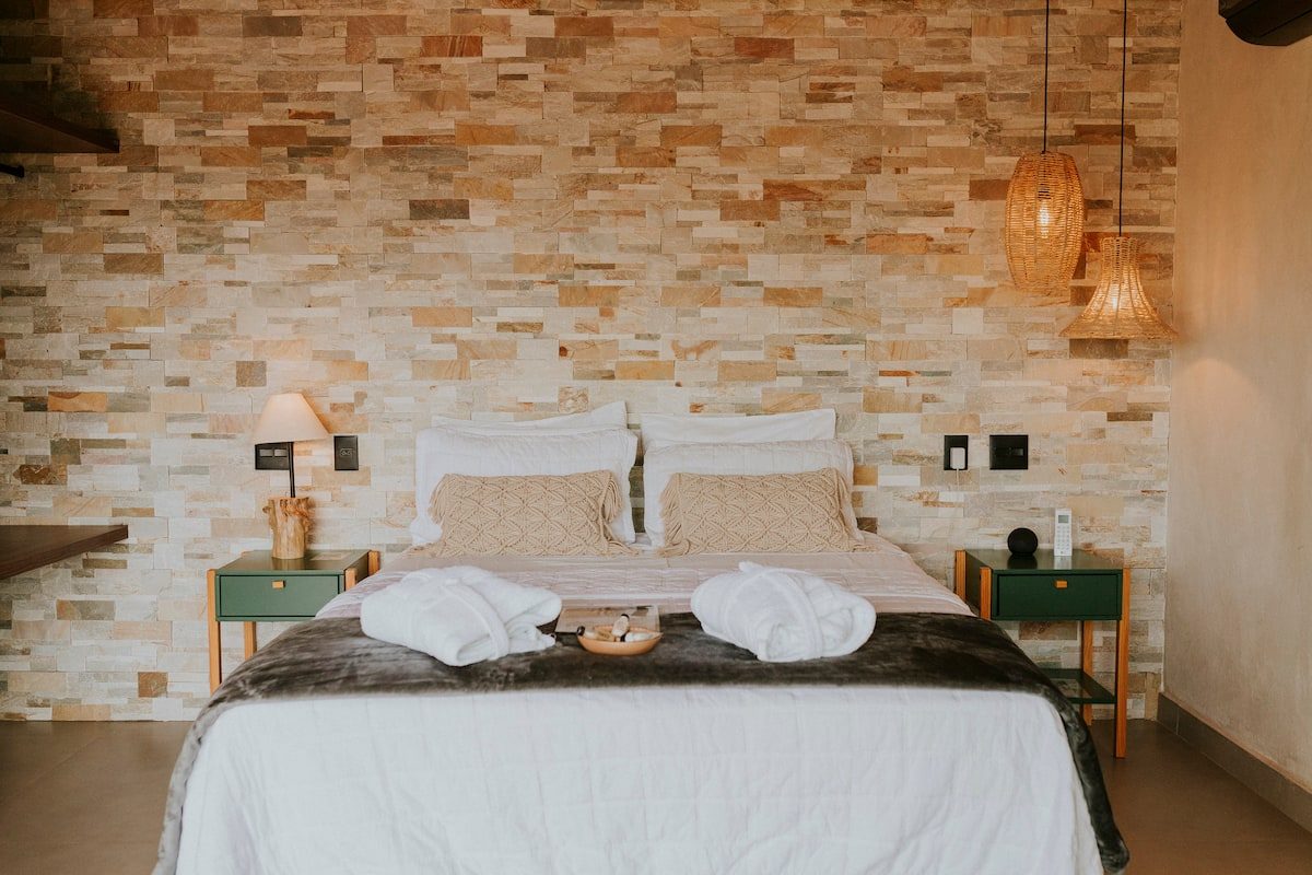 Quarto da Casa Jade com cabeceira de pedra natural, roupões brancos sobre a cama e luminárias de palha