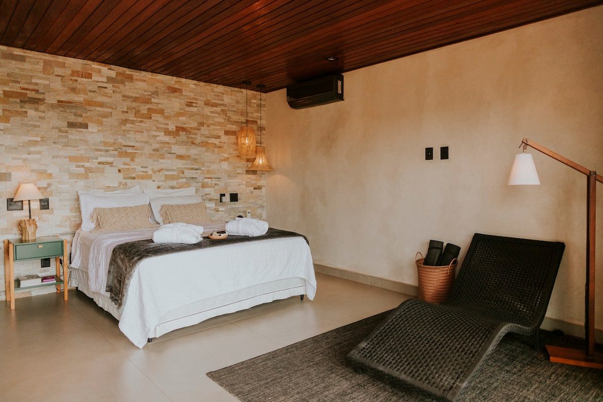 Quarto amplo da Casa Jade com espreguiçadeira de fibra, parede de pedra e cama arrumada com roupões