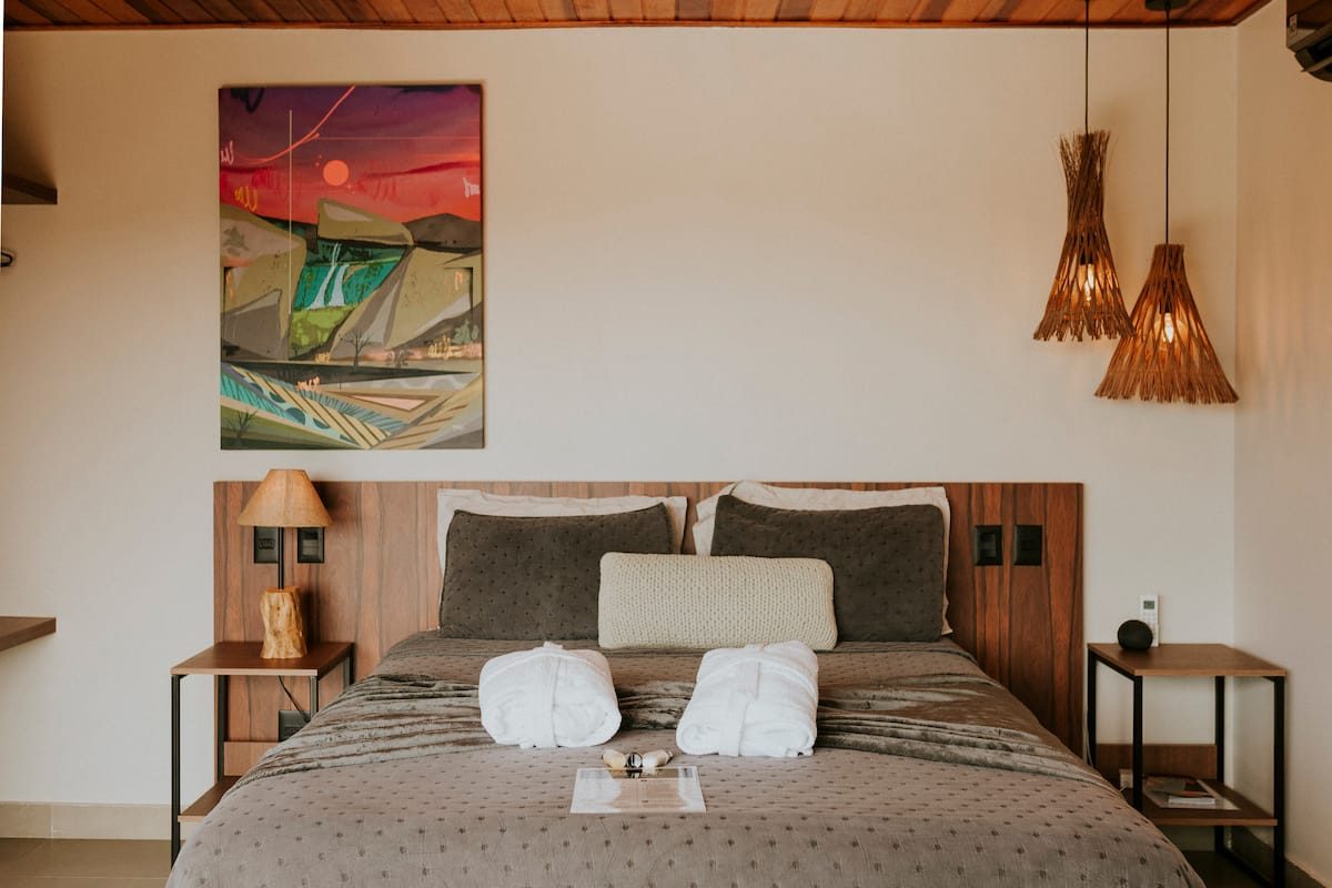 Quarto da Casa Turmalina com cabeceira de madeira, roupões sobre a cama e quadro colorido na parede