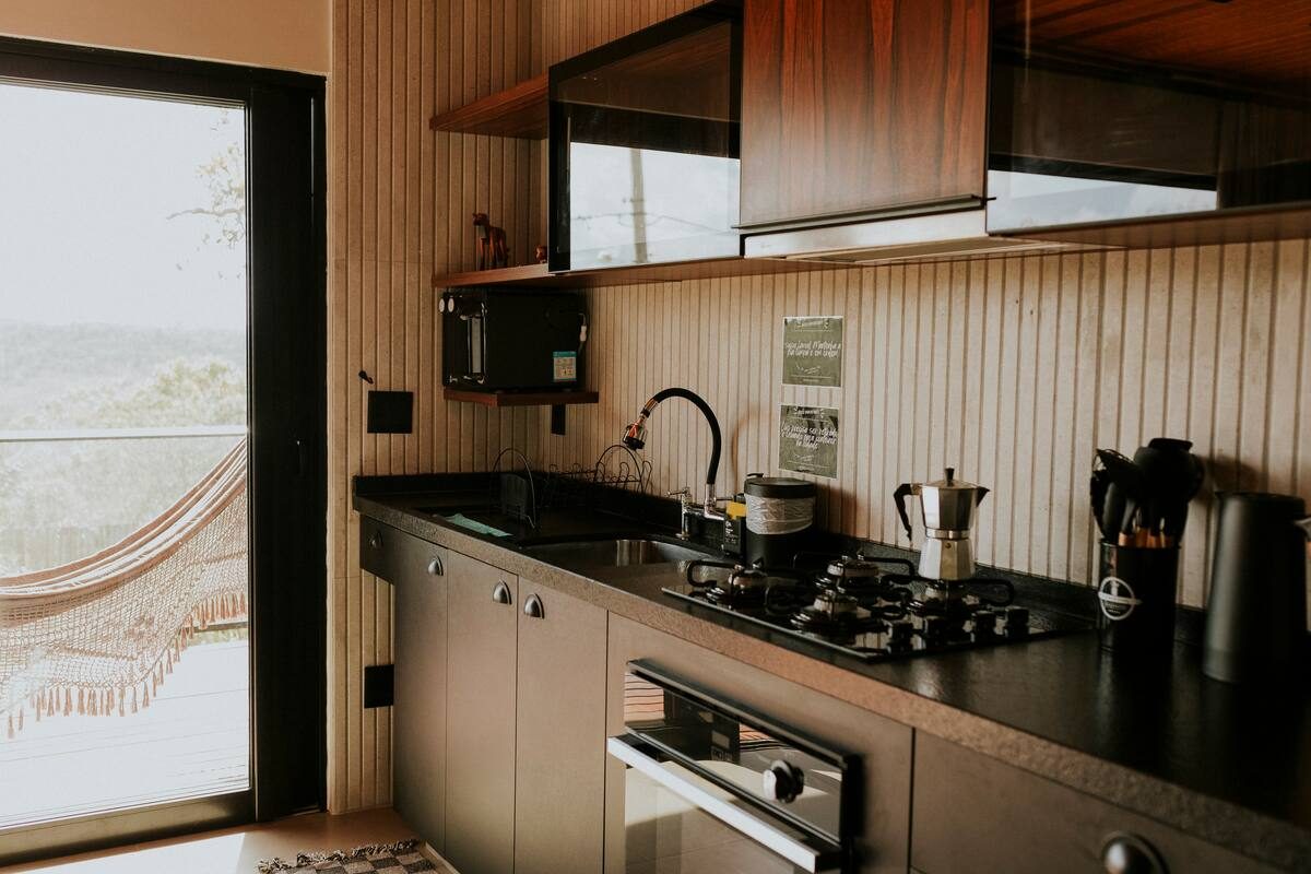 Bancada da cozinha com cafeteira italiana no fogão e a porta de vidro aberta para a varanda com rede